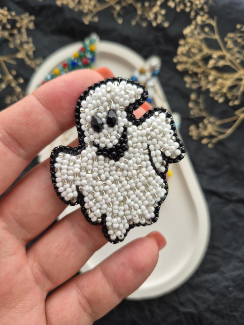 Close-up shot of a handmade ghost brooch embroidered with matte white beads and outlined in black beads. The ghost has large black eyes and a black mouth area. The brooch is resting on the palm of the hand.