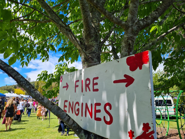 You are looking at a white sign propped in the fork of a green leafy tree on a very sunny day. The sign is hand painted wood with red words saying: "Fire Engines". There are two arrows either side of these words pointing towards the fire engines, which are not visible, but you feel reassured because there's a sign. This is most important, because as anyone who has ever attended a rural fair knows, the fire engines are the coolest part of the whole thing and everyone wants to know where they are urgently. Beyond the sign you can see a big blue sky, green lush grass, and numerous people all wandering around stalls. Most of them walking away from the fire engines, possibly because they have seen the huge queue for them and have decided to get some food first. While looking at this sign, please imagine the sound of well over a thousand people having a very good time, small humans playing. A Morris Dancing's group's penny whistle and accordion and, most importantly of all, the WOOP WOOP noise of the fire engine siren going off in short bursts every now and then when someone is lucky enough to get to press the siren button. The smells in the air are of the nearby Brazilian roast meat truck, the nearby Dutch pancake stall that is currently brewing a very strong batch of coffee in traditional kettles, very green grass and sunscreen. You know, looking at this picture, that if you were here, you would be having a rather marvellous day.