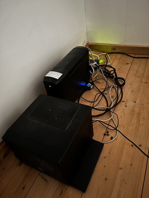 A corner of a room featuring two black electronic devices on a wooden floor. Numerous cables are visible, some leading to a power outlet. One device has a control panel with a blue light.