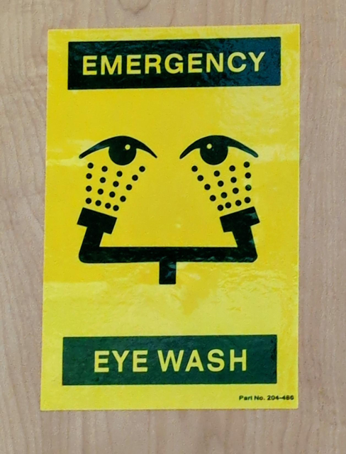 A yellow sign with black lettering that reads "Emergency Eye Wash". It has two half-open eyes with a two-headed shower shooting up into them. The washing mechanism looks like a forced smile, leading into epic eye bags.