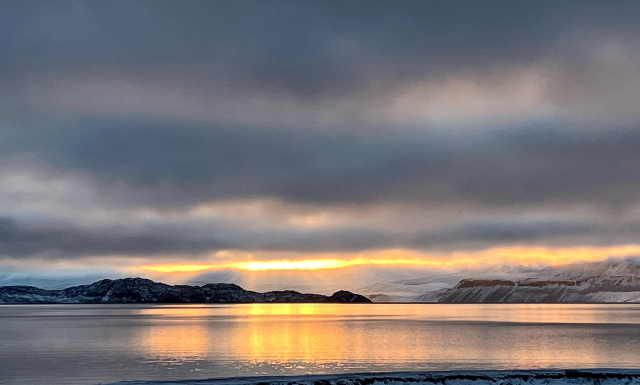 The Sun, low on the horizon at noon, casts a golden light through a break in the clouds. The calm ocean is painted gold by that light. Crepuscular rays can be seen over a black volcanic point. 