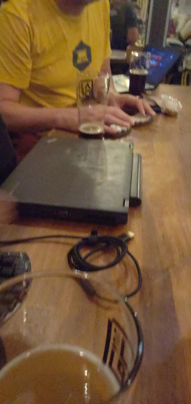Une table de bar avec deux thinkpads et deux claviers ergonomiques, ainsi que plusieurs verres de bierre. La photo est légèrement floue, indiquant le niveau d'alcoolémie des participants