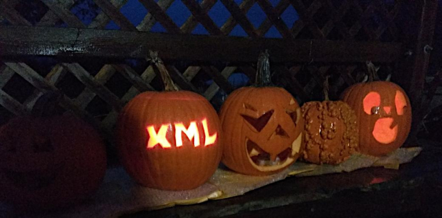 Four carved pumpkins in a row;l the nearest has the letters X M L carved into it, lit up from a candle inside.