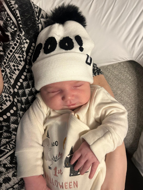 Bertie, all snug and snoozing in festive Halloween style! He's wearing an adorable white beanie with "BOO!" in bold black letters and a cute pom-pom on top. His cozy onesie features playful Halloween graphics, including a ghost, spider web, and a black cat, with the inscription "My 1st Halloween." 