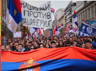 Huge crowds marching with placards and banners march behind a banner made of the Serbian flag