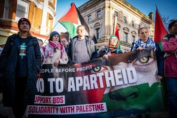 Crowd with flags standing behind a huge banner reading PEOPLE BEFORE PROFIT, NO TO APARTHEID, SUPPORT BDS, SOLIDARITY WITH PALESTINE