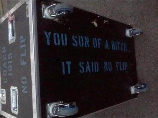 a large rectanguar black road case used for stage productions, tipped onto its side so its stood long ways up. the side facing up says ' coach 1915 no flip' and the bottom, which is now its side exposes four caster wheels and in large text on the underside reads 'you son of a bich it said no flip'