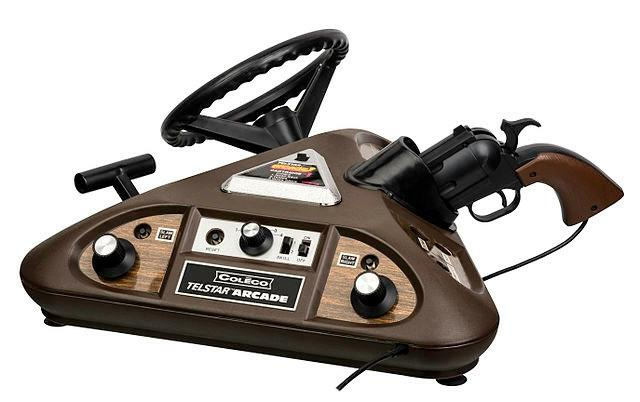 A brown triangular prism. The top side is used to for the triangular top-loading cartridges (this cartridge has Road Race, Tennis, and Quick Draw). The facing side is the pinball interface, with two paddles, a rotary switch, 'Slam Left', 'Slam Right', a skill selector, a power switch, and a reset button. The far side is a racing interface, with steering wheel and gear selector. The right side is shooting game interface, with a realistic looking sidearm and three buttons in holes. The interface panels alternate woodgrain and faux metal.