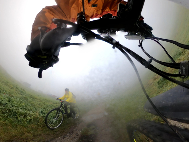 A fisheye view from a handlebar mounted camera, where two cyclist s are passing by on a dirty path in foggy weather. 
