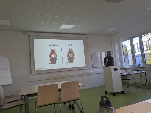 Photo from inside a seminar room. A projected image on the wall is showing a brown Labubu on the left with the text "Labubu" above it, and to the right of it, a second, spikier Labubu with the text "Lakiki" above it. The lecturer is standing at a podium next to the image. He looks as though he is speaking and explaining a complex subject.