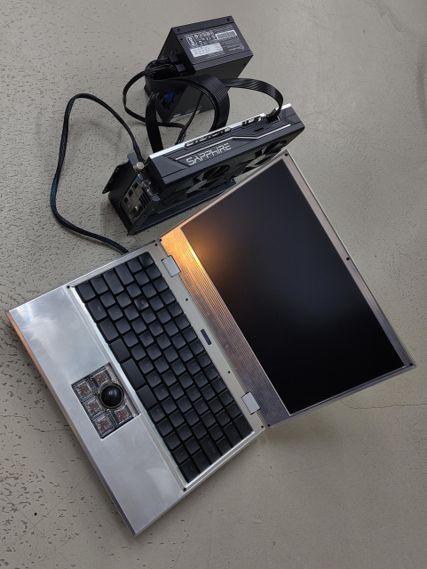 a silver colored raw aluminum laptop with a black/transparent trackball sits on a concrete floor. it's connected with a cable to an amd gpu that is connected to an sfx power supply with black cabling, all items sitting on the ground