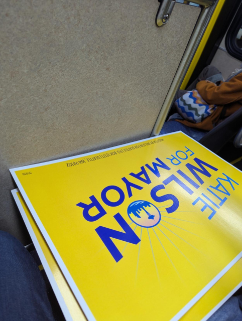 Two posters on the lap next to me that say Katie Wilson for Mayor.