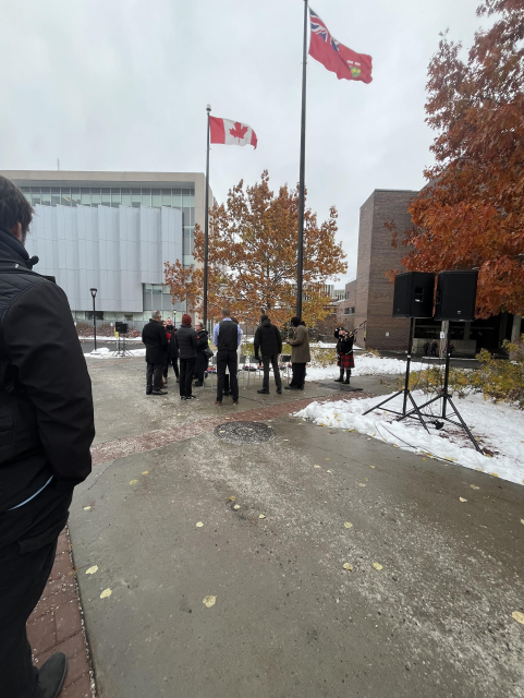 A small group of people in uniform stand near a Canadian and Ontario flag outside some buildings. There is are some maple trees turning rusty red. There is a small amount of snow on the ground and the path looks wet with salted melt.  There is a bagpiper.