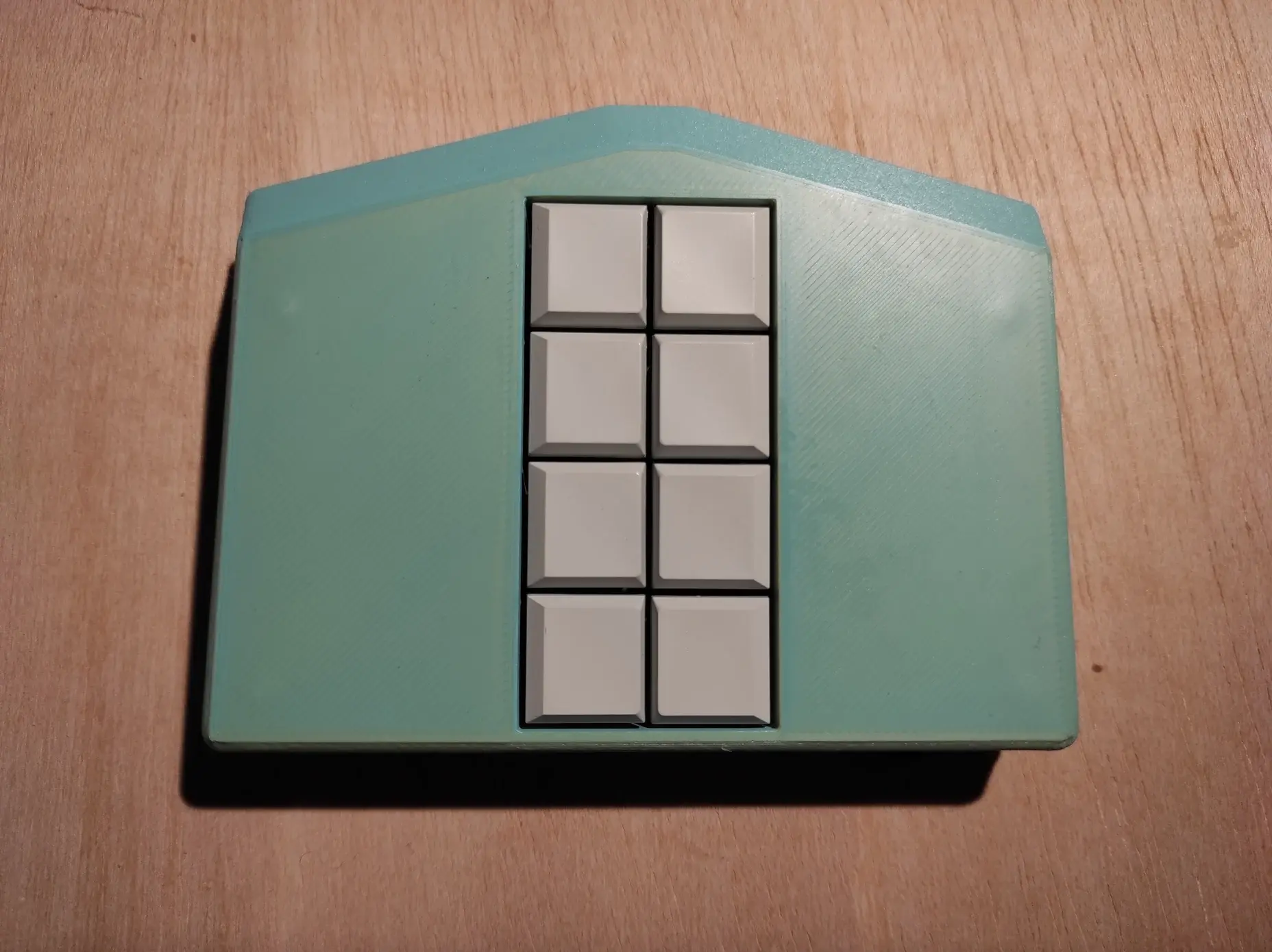 Mobile Braille-Tastatur Metabraille mit 8 sichtbaren Tasten. Das Gehäuse ist türkis und die Tasten sind weiß. Das Gehäuse ist ungefähr fünfeckig. Ein Rechteck wird an der Breitseite durch ein Trapez erweitert. Die Form ähnelt einem Haus mit Fenstern in der Mitte.