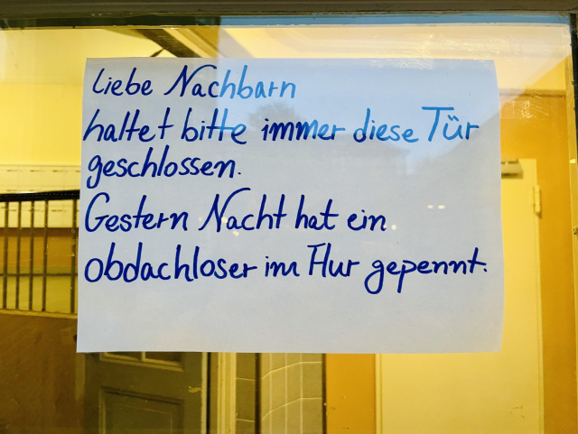 Ein handgeschriebener Zettel an eine Haustür geklebt: Liebe Nachbarn, haltet bitte immer diese Tür geschlossen. Gestern Nacht hat ein Obdachloser im Flur gepennt. 