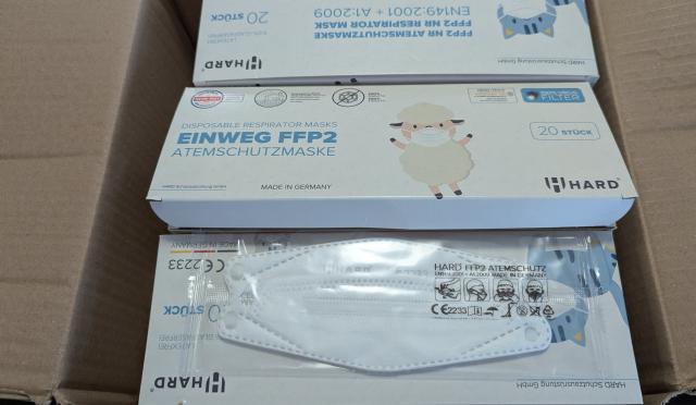 Eine andere Ansicht des Kartons, daneben eine aus den Karton ausgepackt, aber noch in der Schutzfolie eingeschweißte Maske.
