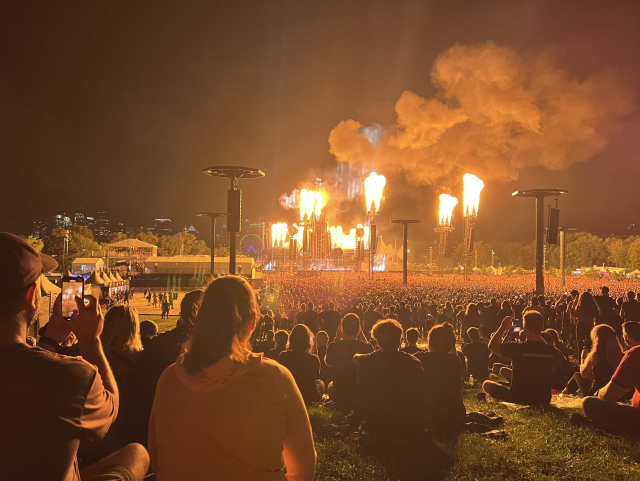 AI, The image displays a large outdoor concert at night, with a massive crowd gathered in front of a stage. Bright flames shoot up from the stage, illuminating the scene. In the foreground, people are sitting on the grass, watching the performance, with some taking pictures or videos on their phones. The atmosphere seems lively and energetic, with a backdrop of city lights visible in the distance. 

