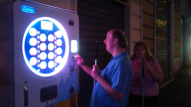 AI, A man is using a large vending machine with a bright, circular display that has the Durex brand visible on it. The vending machine is on the side of a street or closed shop. The man is holding a white cane, and a woman standing behind him is also holding a white cane, indicating they may be visually impaired. The scene is dimly lit, with the vending machine illuminated in contrast to the dark surroundings.

