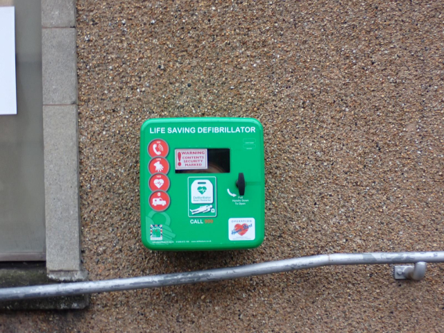 A green defibrillator box mounted on a harmed or pebbledash wall.