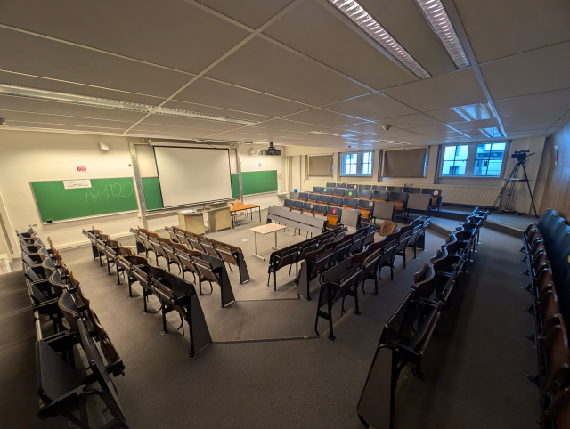 An empty lecture room with tiered seating arranged in a horseshoe around a central stage and projector screen. Taken before the Geospatial devroom began, the room later filled up with a full audience and a queue outside.