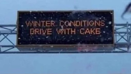 Overhead electronic road sign reads "WINTER CONDITIONS, DRIVE WITH CAKE."