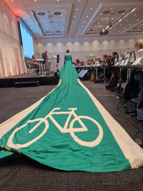 A woman seen from behind in a green dress with a 10+ meter long train behind it. The train is outlined in thick white with a bicycle symbol, representing a cycle lane