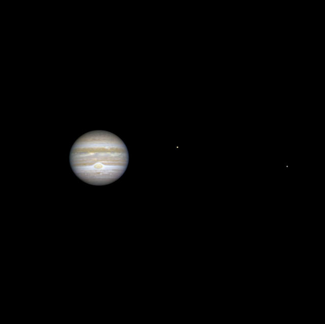 Image captured by the Mars Reconnaissance Orbiter showing Jupiter and two of its moons. Jupiter appears slightly wider than tall, with orange and white atmospheric bands and a large oval orange storm below the center. Two small bright spheres to the right are its satellites.