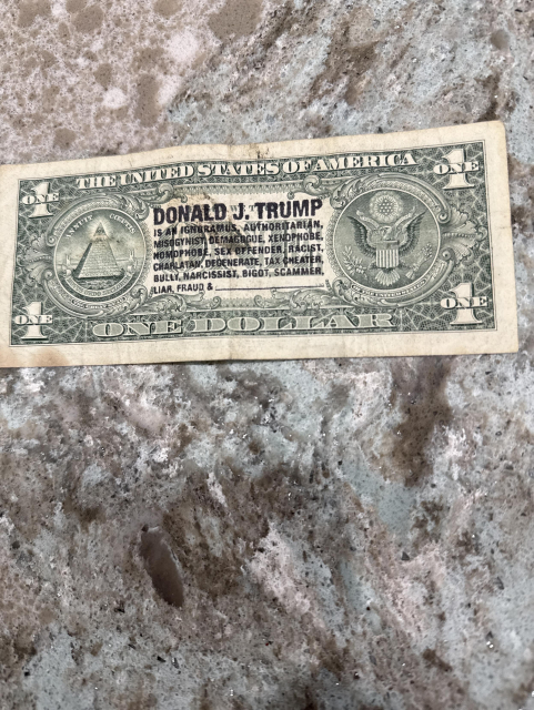 A worn U.S. one-dollar bill with additional printed text in black ink over the standard design. The bill features the standard symbols of the pyramid with the eye and the Great Seal, but in the center, large text reads "DONALD J. TRUMP" followed by several derogatory terms and accusations. The printed text is as follows:

"**DONALD J. TRUMP**
IS AN IGNORAMUS, AUTHORITARIAN,
MISOGYNIST, DEMAGOGUE, XENOPHOBE,
MORONPHOBE, SEX OFFENDER, RACIST,
CHARLATAN, DEGENERATE, TAX CHEATER,
BULLY, NARCISSIST, BIGOT, SCAMMER,
LIAR, FRAUD &"

The extra text appears to be meant as a strong political statement against Donald Trump, stamped directly over the original dollar bill design. The rest of the bill keeps its standard appearance. If you need more details or want me to transcribe anything else, please let me know.

Image description provided with Be My Eyes.