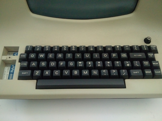 A close-up picture of an ADM 3a terminal keyboard. The left arrow label is above the "H" key, the down arrow label is above the "J" key, the up arrow label is above the "K" key, and the right arrow label is above the "L" key. In the top right, a single key is labeled with a circumflex, a tilde, and the word "HOME".