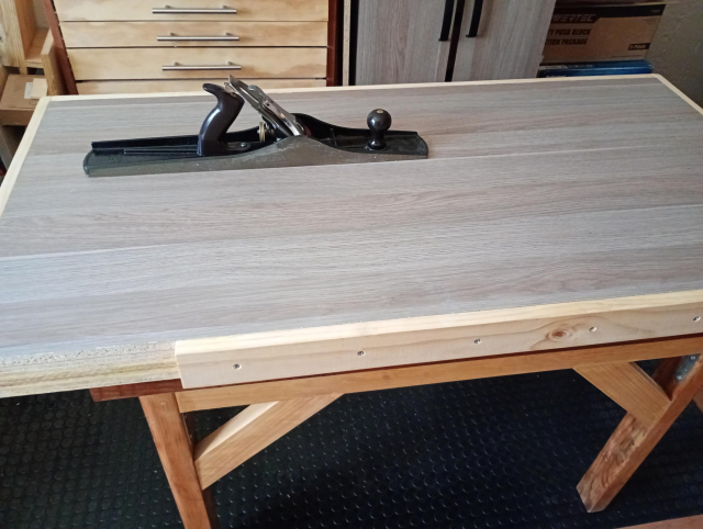 La tapa del banco de trabajo, 3 hojas de triplay de 18mm más 1 hoja de melamina de 15mm, enmarcadas con pino de 1x3 (menos dos tramos para las prensas). Descansa sobre mi mesa restaurada y reforzada. Sobre el banco, un cepillo Stanley no. 7 de 50cm de largo.