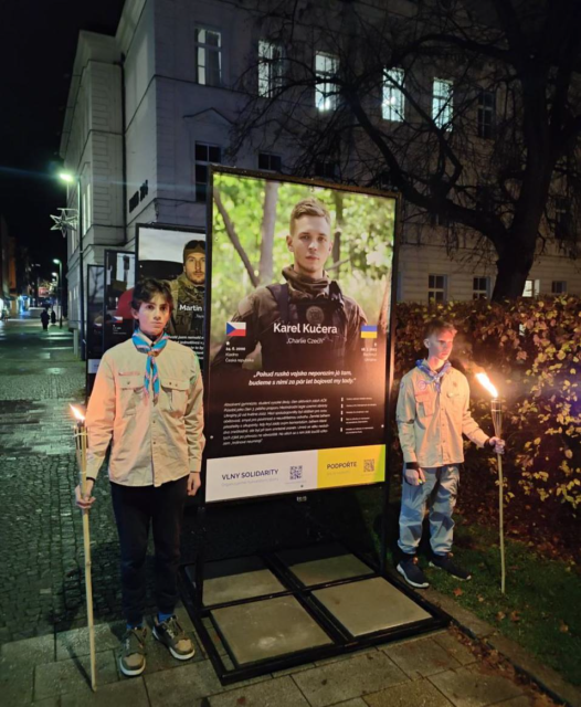 Young people standing next to a banner with portrait of Karel Kucera with his biography on an evening street