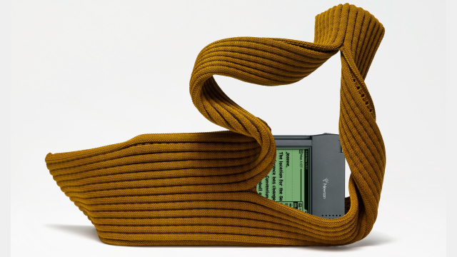 An artistic representation of a mustard-colored, textured fabric that is twisted and shaped, partially enclosing a vintage Apple Newton MessagePad 2000 with a monochrome green screen UX elements. The background is plain white, emphasizing the unique design.