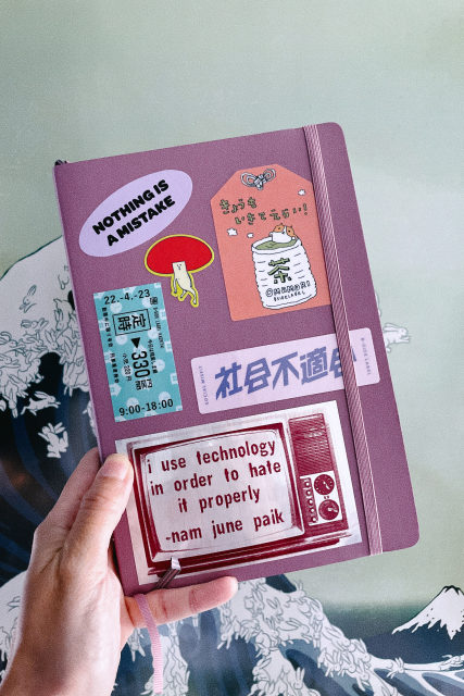 A hand holding a dusty pink notebook covered with stickers, clockwise: 1. A purple oval with the words “Nothing is a mistake”, 2. A sort of mushroom character striking a pose, 3. A lucky charm-like sticker with an illustration of a hamster sitting in a teacup of green tea and in Japanese says “I lived today, too!”, 4. A pink rectangle with Japanese kanji character for “social misfit”, 5. An old TV set with a quote by Nam June Paik: “I use technology in order to hate it properly.”, 6. A sticker that copies the look of a Japanese train ticket. 