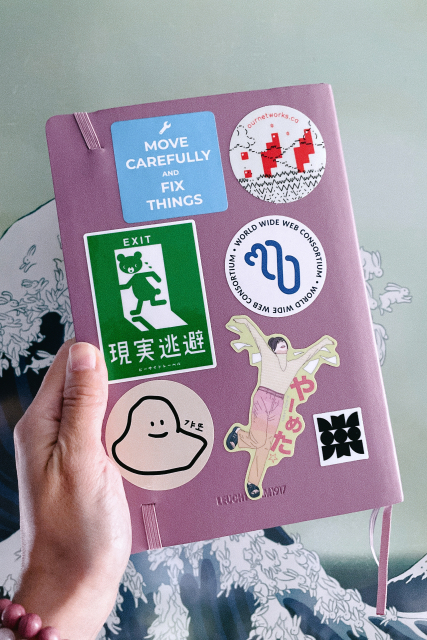 The back of the same notebook, also covered in stickers, clockwise: 1. A blue square with an icon of a wrench and the words “Move carefully and fix things”, 2. Ascii art of mountains, clouds, and grass and the Our Networks logo pixelised, 3. The W3C (World Wide Web Consortium)’s new logo (it is upside down), 3. The Merveilles Mastodon community’s logo, 4. A femme person jumping for joy with the words in Japanese “I quit (my job)!”, 5. The mascot for a local restaurant named Gateau Ghost, it is an illustration of some sort of  blob character with eyes and a smile, 6. A spoof of the green exit sign, except this time a bear is the one running out the door. There are Japanese characters underneath it that (I think) mean “Exit” or “Escape”.
