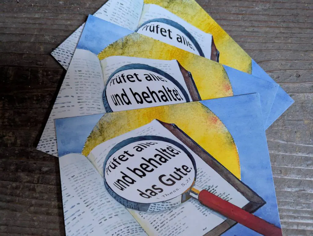 Auf einer Holztreppe liegen Postkarten. Eine Lupe ist darauf abgebildet, die den Satz vergrößert: Prüfet alles und behaltet das Gute.