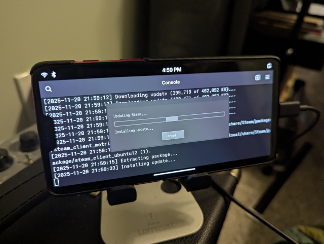 Photo of a phone on a stand running PostmarketOS, displaying a terminal with a logs and Steam's grey "Updating Steam" window.