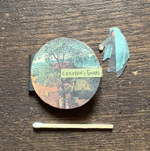 A circular book featuring “Children’s Games” by Pieter Bruegel the Elder. The cover shows an image of trees from the painting. Next to the book is a small cut-out figure in a blue cape from the same painting, placed alongside a matchstick for scale.