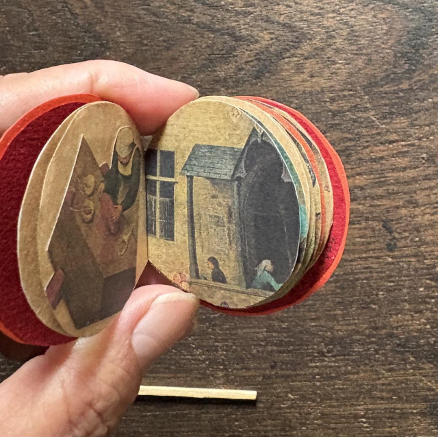 A hand holding the book open to show the inside pages. The pages feature cut-out details from Pieter Bruegel the Elder’s “Children’s Games.”