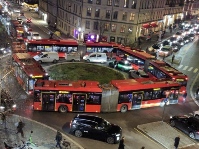 A scene showing four red bendy buses making up a squarw around a small roundabout. There are cars trapped inside them and long lines on the two visible sides going into the roundabout. Photo was found in Avisa Oslo, a local newspaper, and was sent them by a guy named Endre Helgeland. 