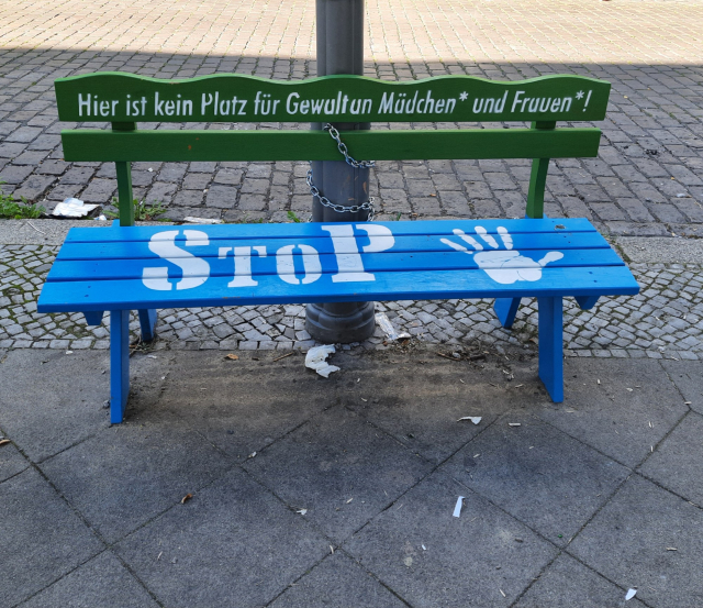 Eine zweifarbige Holzbank im öffentlichen Raum. Die Rückenlehne ist grün und trägt die weiße Aufschrift „Hier ist kein Platz für Gewalt an Mädchen* und Frauen*!“. Die Sitzfläche ist blau und zeigt in Weiß das Wort „STOP“ sowie einen Handabdruck. Die Bank ist mit einer Kette an einem Laternenmast befestigt. Der Boden besteht aus Gehwegplatten und Kopfsteinpflaster.