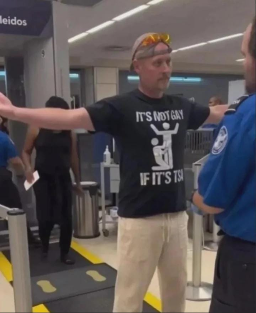 alt : at the Airport Security Check, after scanning, a man spreads arms waiting for the security employee toi pat him.
His illustrated t-shirt says : It's not gay if it's TSA. The line art drawing depicts a patting scene where the employee is on his knees.

