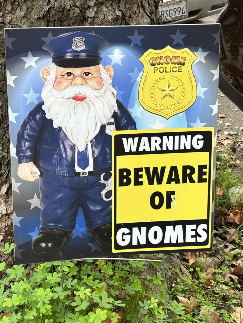 A photo of a “warning beware of gnomes” yard sign featuring a old gnome man in a police uniform with the badge “gnome police”