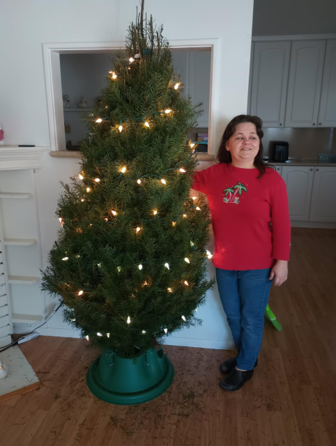 Jessica stands in a red sweatshirt with palm trees and Christmas presents, and blue jeans. The very bushy and full Christmas tree stands beside her, about a head taller than her. Lights are on it but no ornaments yet.