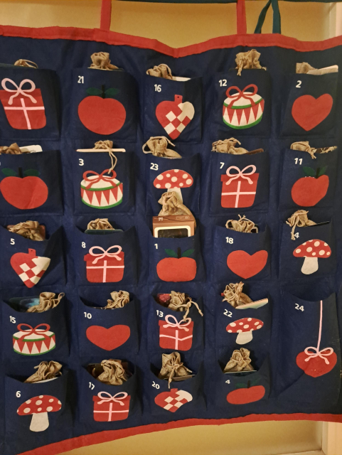 Photo of a square of blue fabric with 24 large pockets. Each pocket has a different red and white design: hearts, gift boxes, mushrooms, apples. Peeking out of the pockets are smaller gift bags and cards.