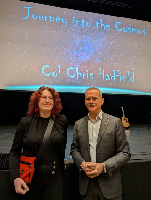 Na fotografii stojí vedle sebe Kayla Eilhart (vlevo) a astronaut Chris Hadfield (vpravo) u pódia. Za nimi svítí velké promítací plátno s hvězdným pozadím.