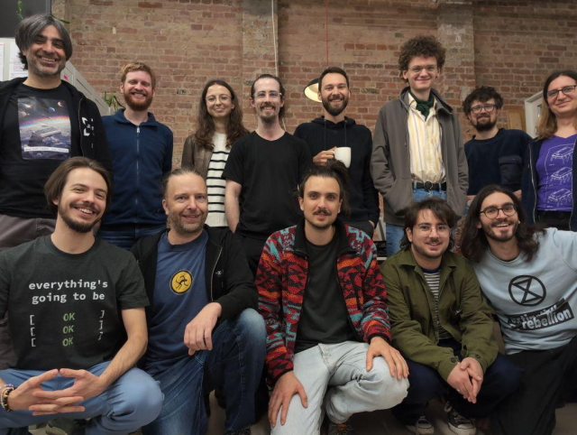 Group photo with 13 hackers in two rows, with a brick wall in the background, inside Cultivation Space.