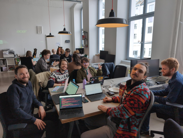 A dozen or so hackers working at 3 tables at Cultivation Space, some of them smiling for the camera.