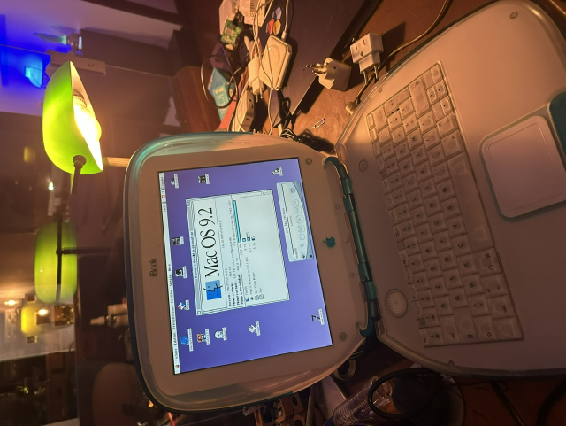 A G3 iBook with an AzERTY keyboard  displaying the OS 9.2 about box on top of a messy table with a green desk lamp in the back reflecting itself into a glass