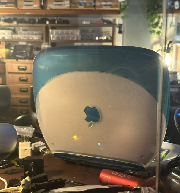 Back of that same iBook G3 seen from behind the glass outside the office. The Apple logo on the open lid is upside down. There’s a lot of mess around.