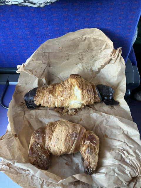 A very flaky chocolate-dipped butter croissant (top) and a plain lard croissant (bottom) on a piece of greaseproof paper. The croissants look a bit battered, and some of the crust has flaked off.